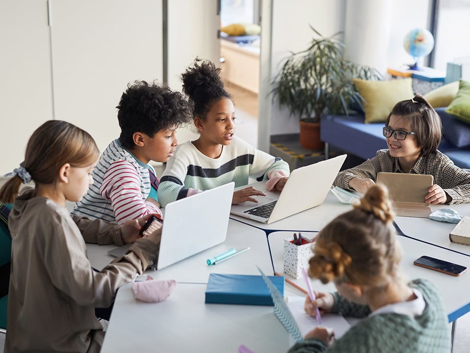 Group of schoolkids sit around table with compuers