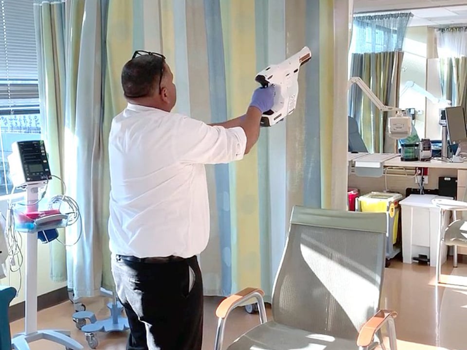 Man wearing gloves holds electrostatic sprayer in hospital room
